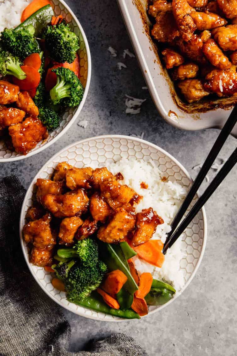 A white bowl of sweet and sour chicken in the bottom left with a pan of sweet and sour chicken in the top right.