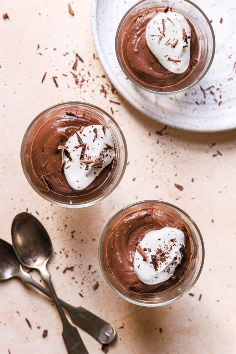 A top down photo with two glasses of pudding topped with whipped cream on the pink counter with two spoons to the left and a white plate with a glass of chocolate pudding on top.