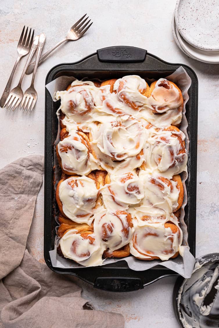 A rectangular baking pan lined with parchment paper with cinnamon roll covered in cream cheese icing on a counter with a stack of plates in the back right.