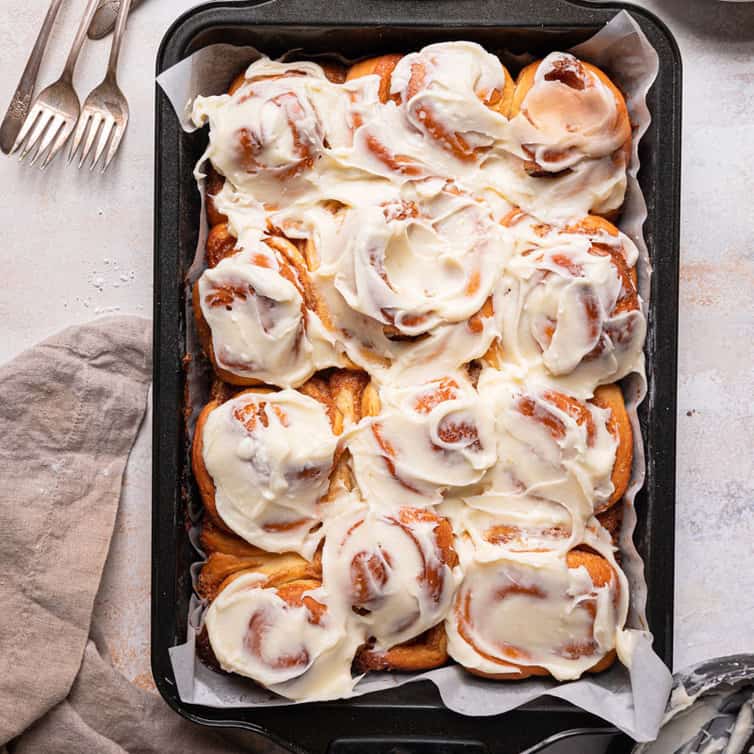 A baking pan with cinnamon rolls covered in cream cheese frosting with a stack of forks in the top left.