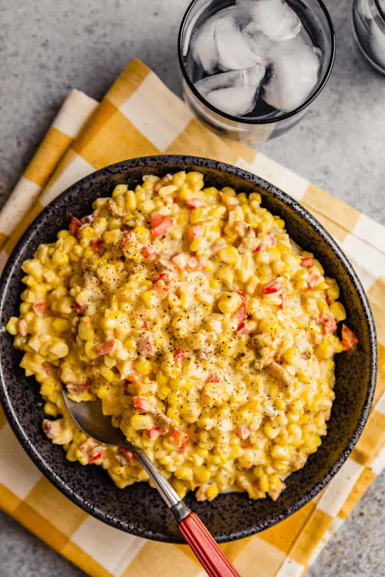 A full black bowl with Mexican corn salad on a yellow and white towel with a glass of water in the top right.