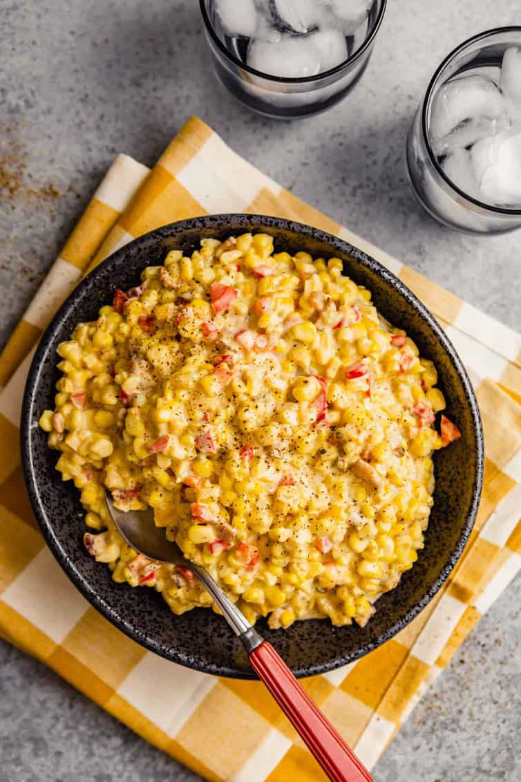 A top down photo of a black bowl filled with Mexican corn salad on a white and yellow towel with two cups of water in the top right.