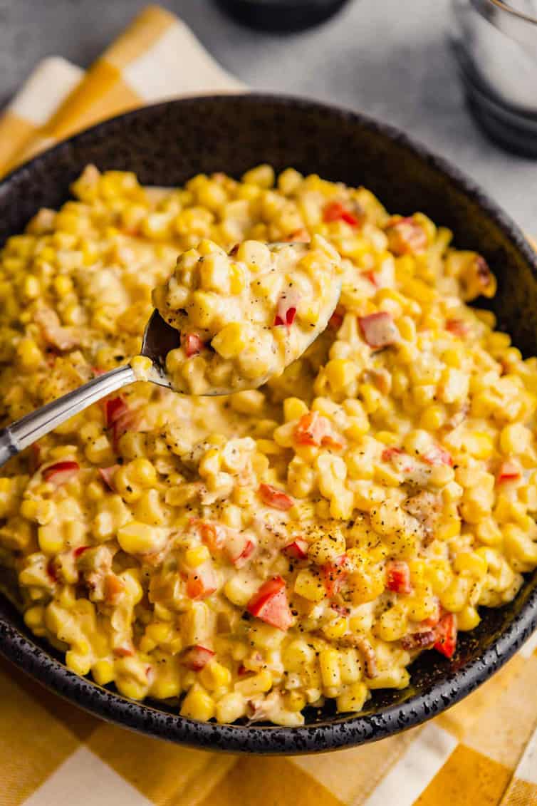 A silver spoon with a scoop of Mexican corn salad over a large bowl of corn salad on a yellow and white towel.