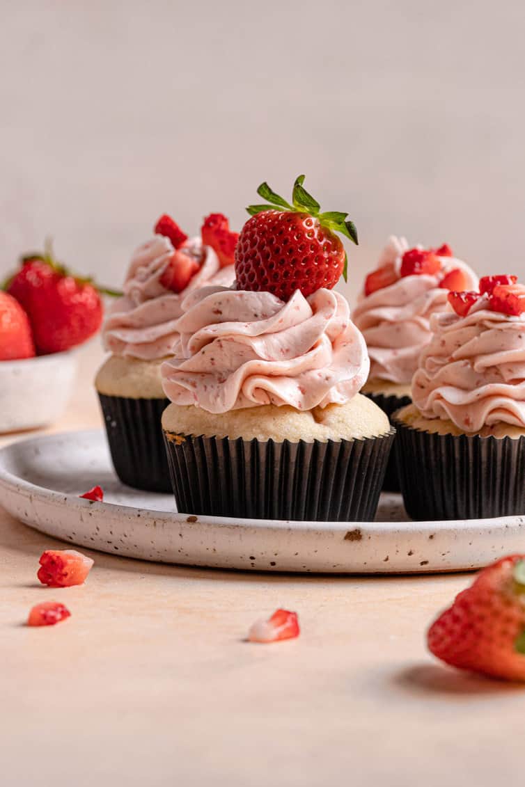 A white plate with a rim holding four strawberry cupcakes topped with fresh strawberries on a pink counter.