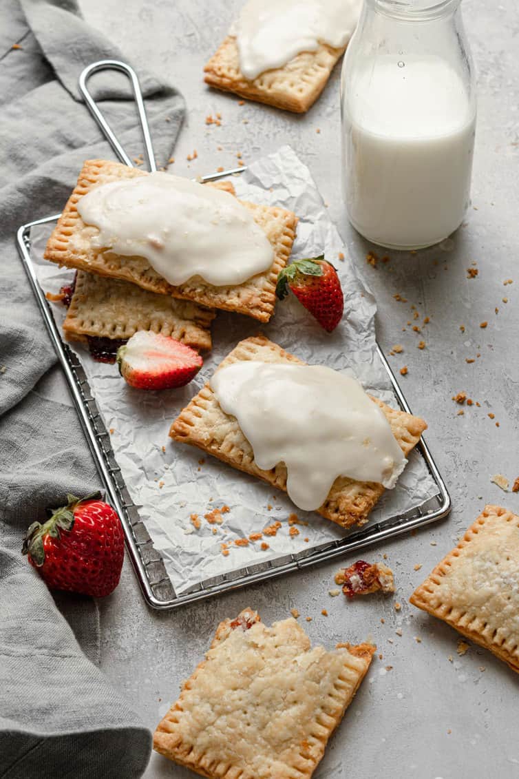 Iced homemade strawberry pop tarts on a baking sheet with parchment paper with a glass of milk to the right and fresh strawberries around.