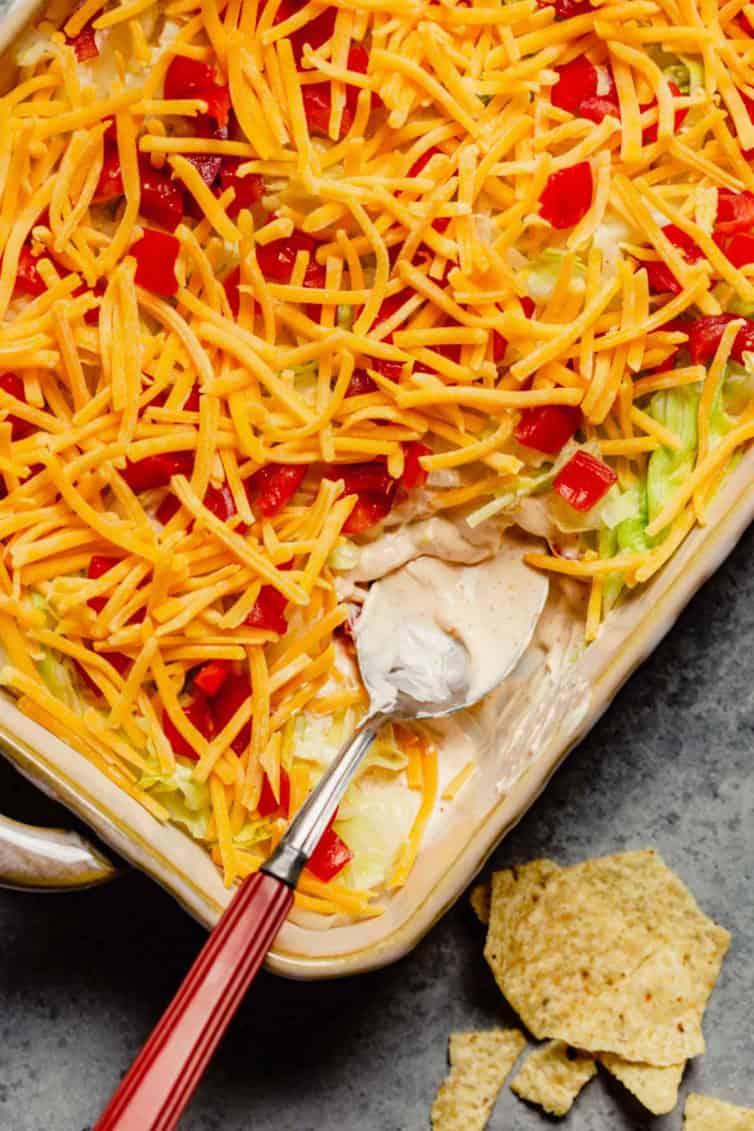 A spoon with a scoop out of the taco dip dish with a few tortilla chips in the bottom right corner.