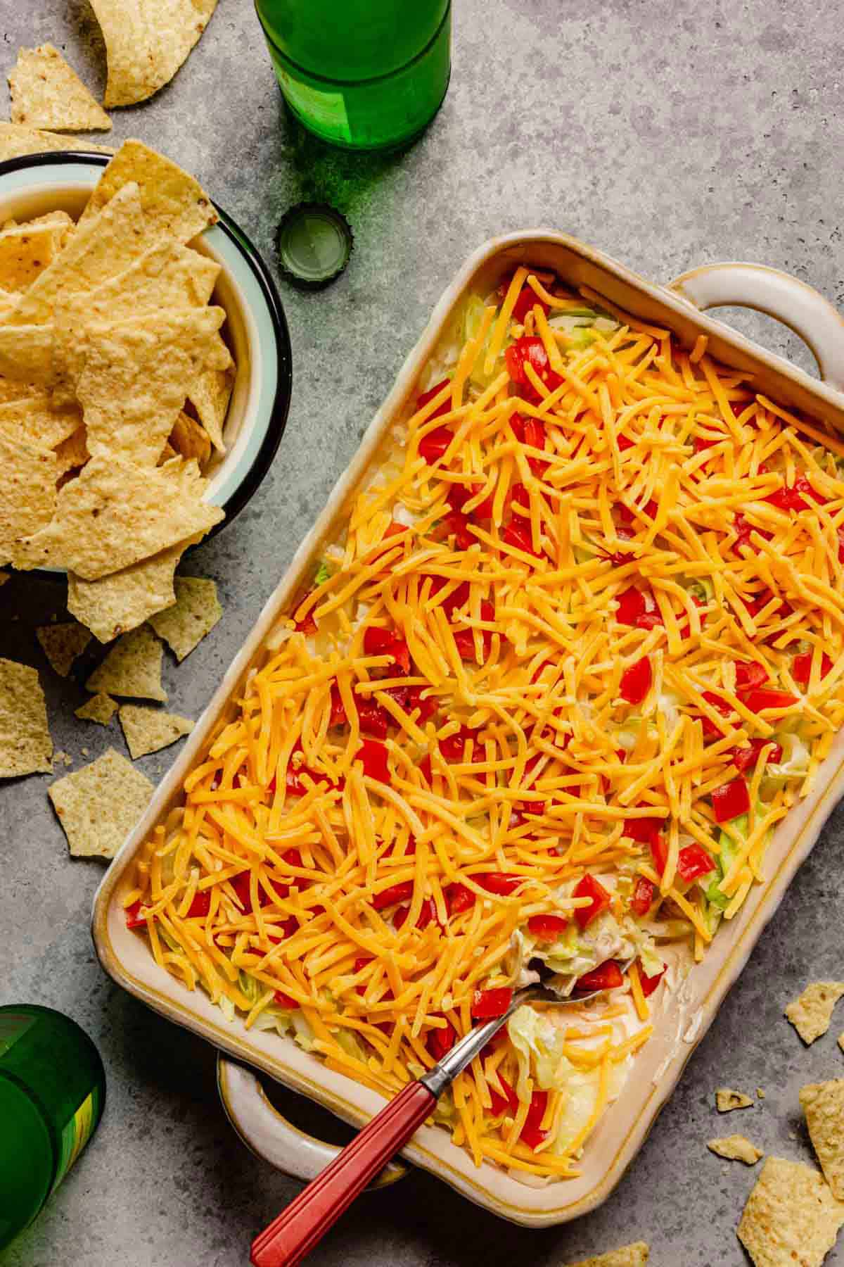 Cheesy no-bake taco dip in a casserole dish with a spoon in the right corner of the dip and a bowl of tortilla chips to the left of the dip.
