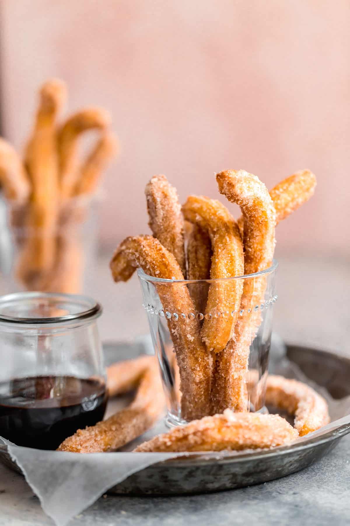 A pie pan with parchment paper with churros in a glass jar on the left and chocolate dipping sauce on the right.