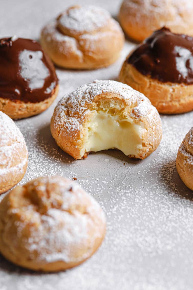 Cream puffs with powdered sugar and chocolate glaze on a counter with one in the middle missing a bite.
