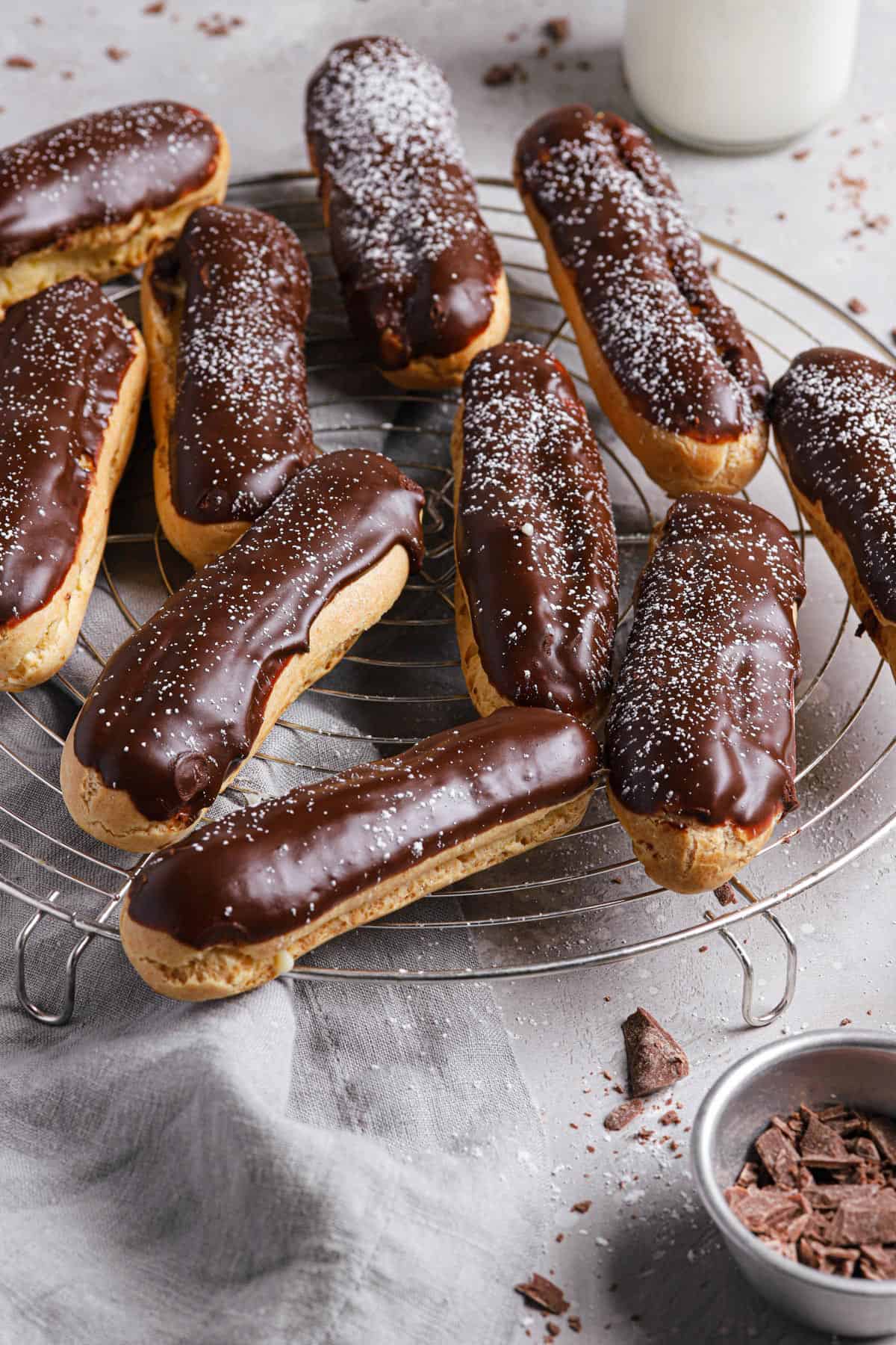 A bowl of cocoa powder in the front right with a wire cooling rack of eclairs topped with chocolate glaze and dusted with powdered sugar.