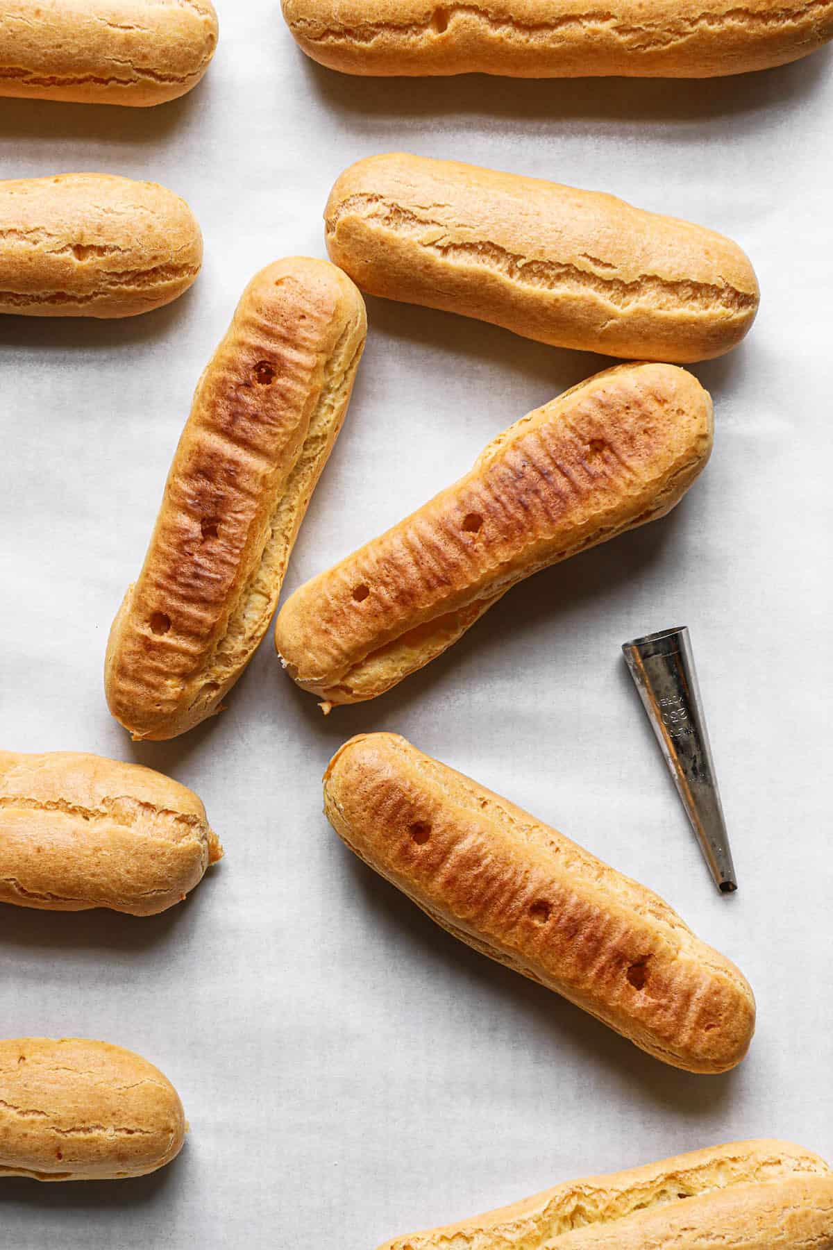 Empty eclair shells on a piece of parchment paper with holes poked in the bottom and a piping tip to the right.