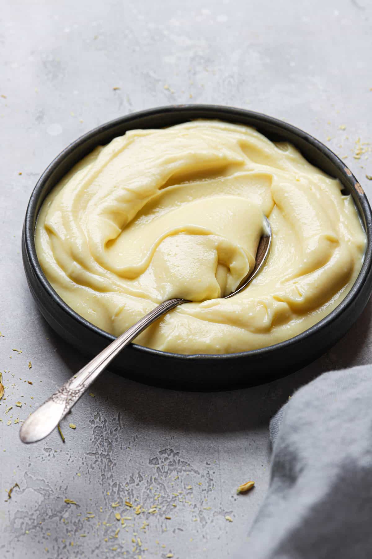 A black bowl of pastry cream with a spoon in the left side of the bowl on a grey counter with a light grey towel.