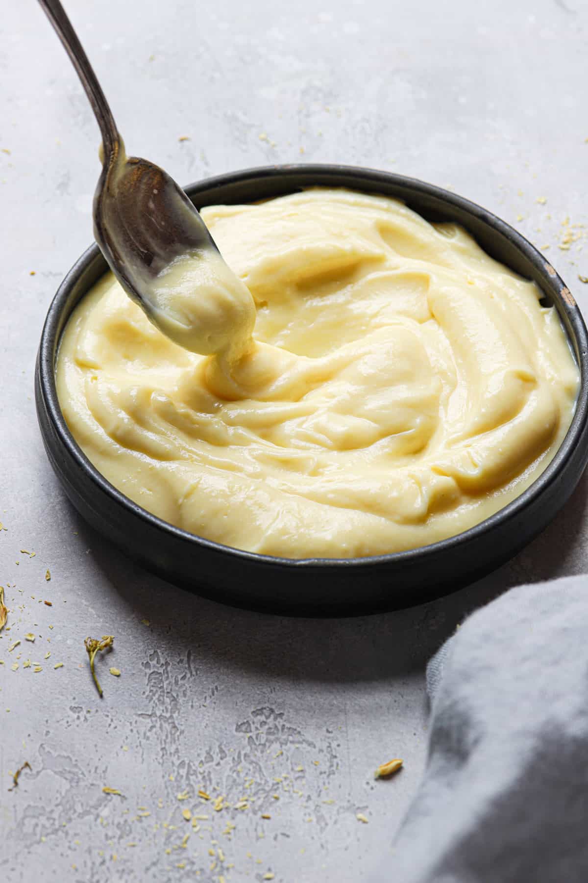 A black bowl of vanilla custard with a spoon being held over the left side of pastry cream on a grey counter with a light grey towel in the bottom right corner.