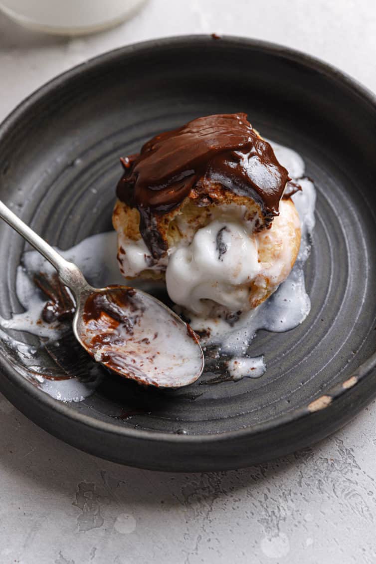 A dark grey plate with an ice cream filled profiterole topped with chocolate sauce and a spoon to the left.
