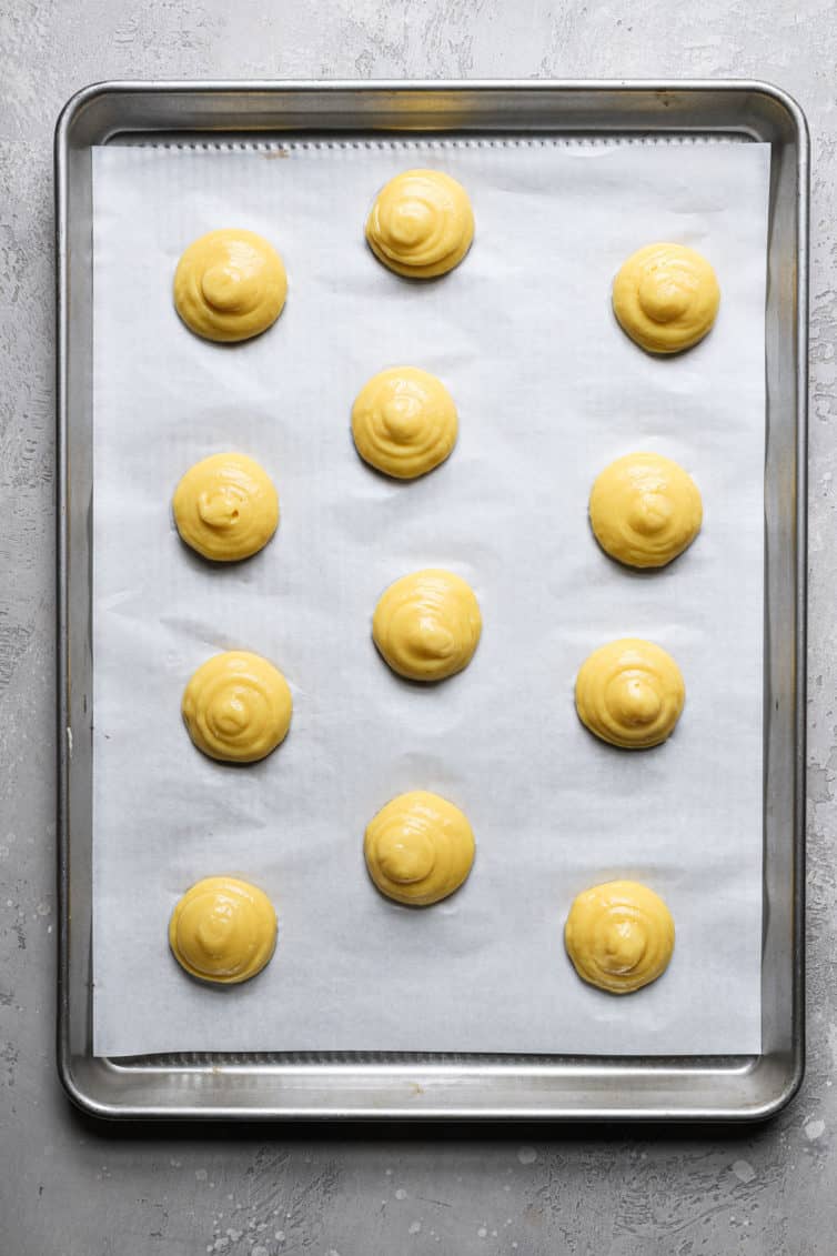 A rimmed baking sheet with a piece of parchment paper with round choux pastry puffs before baking.