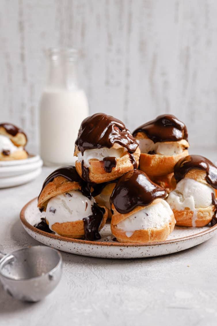A white plate with a tower of profiteroles topped with chocolate and an ice cream scoop in the front.