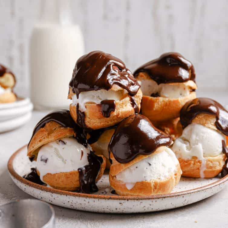 A white plate with a stack of profiteroles topped with melted chocolate sauce and a jar of milk in the back.