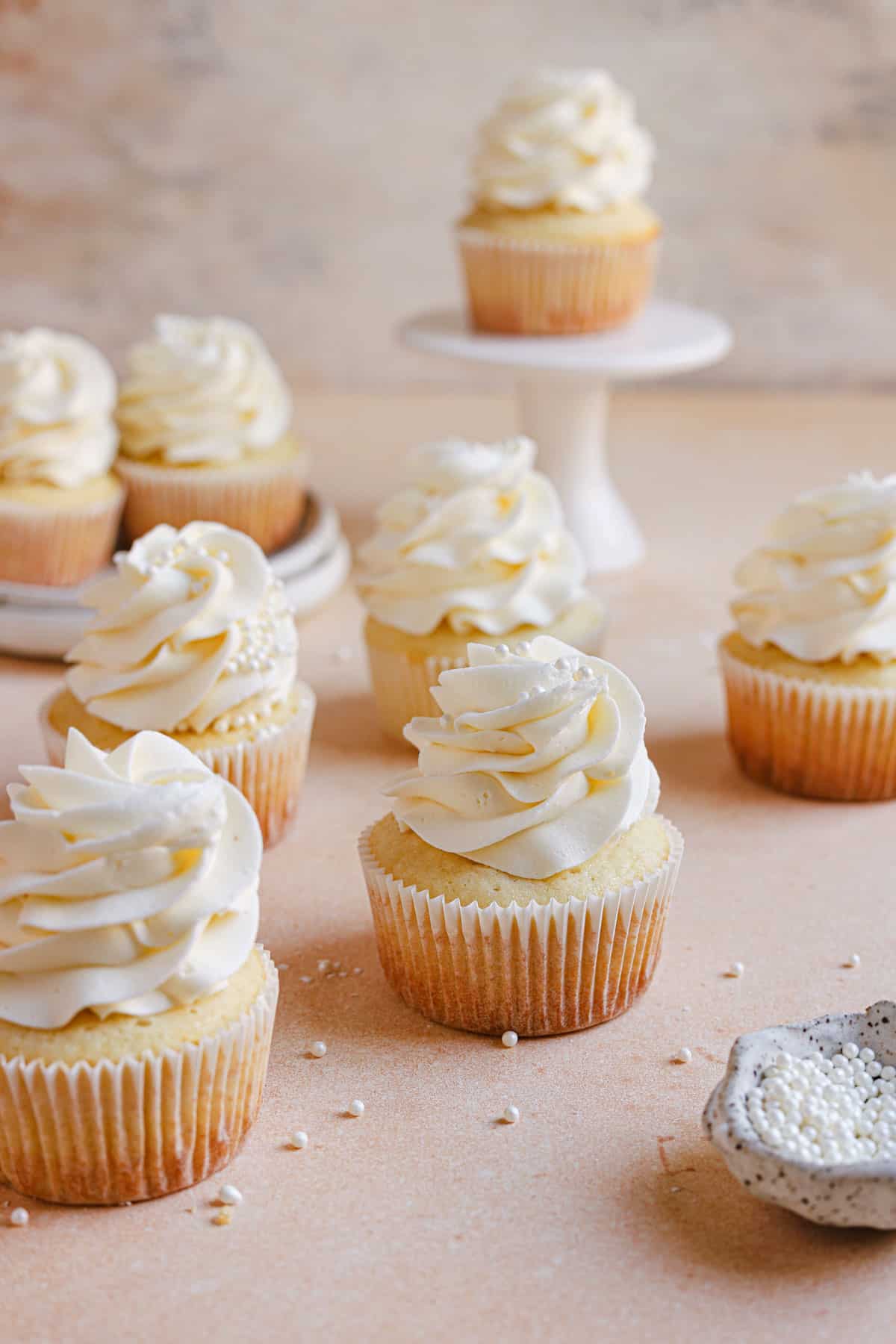 Moist vanilla cupcakes on a counter with a cupcake on a cake stand in the back.