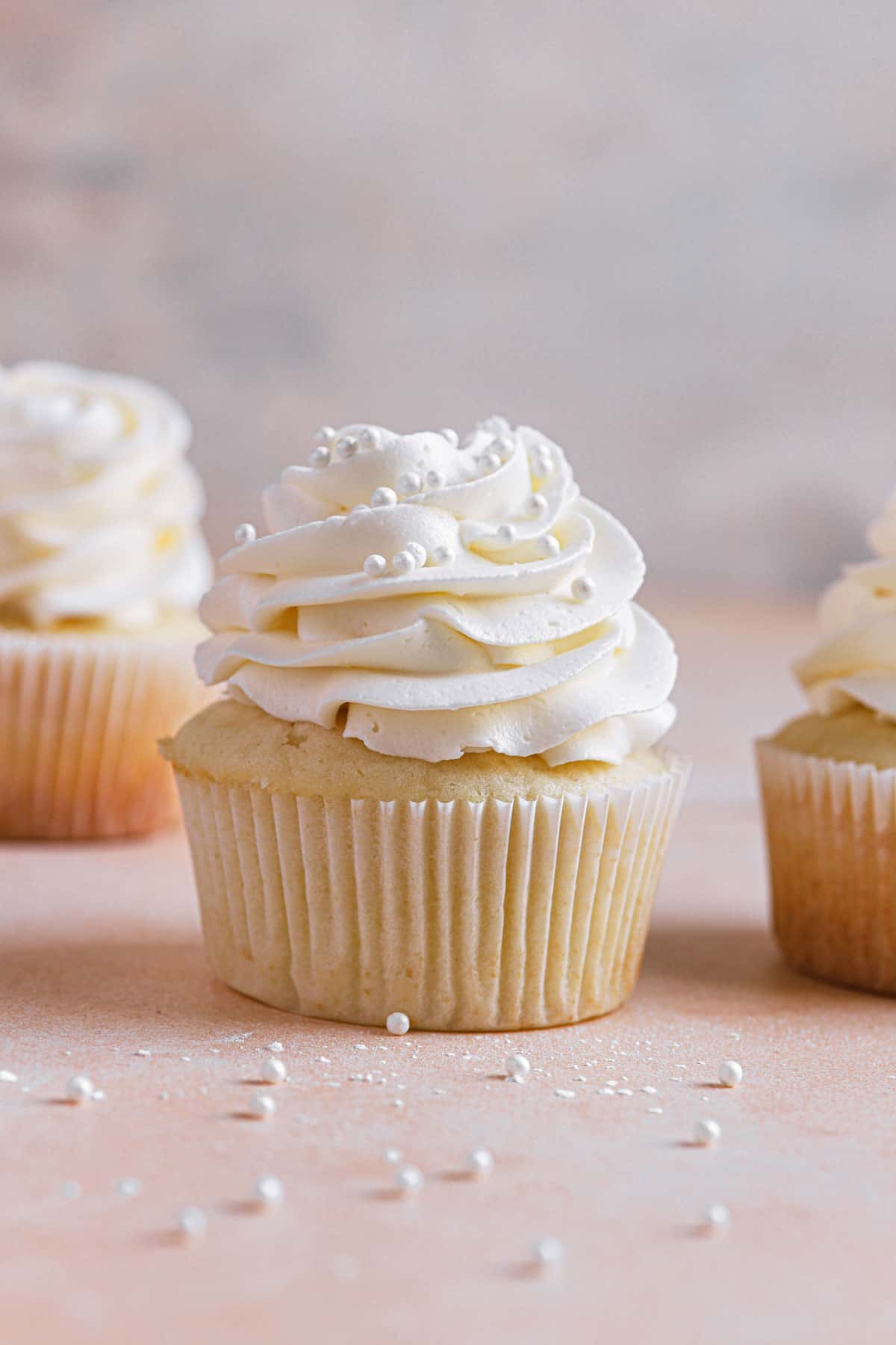 A vanilla cupcake with vanilla frosting and sprinkles in the middle another to the right and one in the back left.