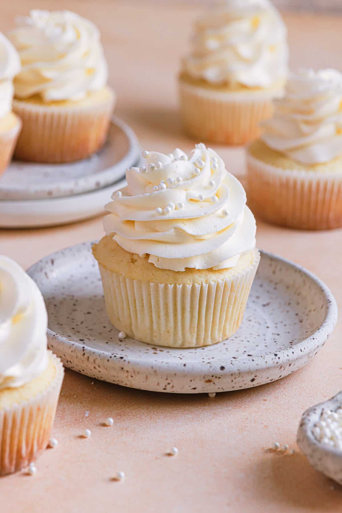 A white speckled plate with a vanilla cupcake in the center and other vanilla cupcakes on the counter in the back.