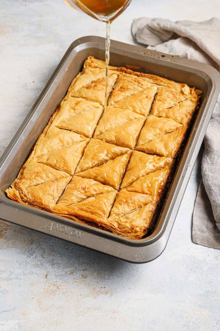 Syrup being poured over a baking pan of baklava with a towel in the back right.