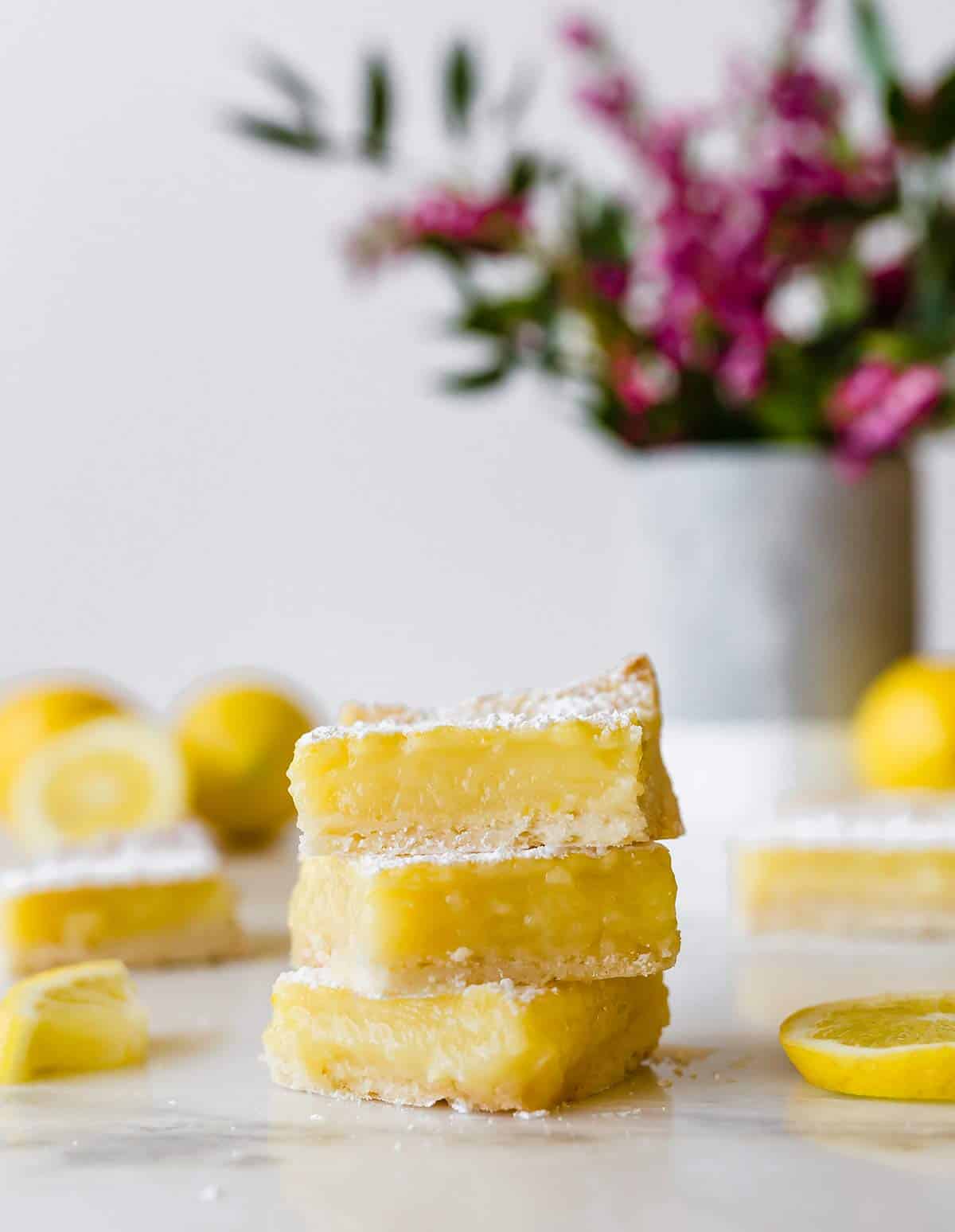A stack of three lemon bars with cut lemons and a vase of flowers in the background.