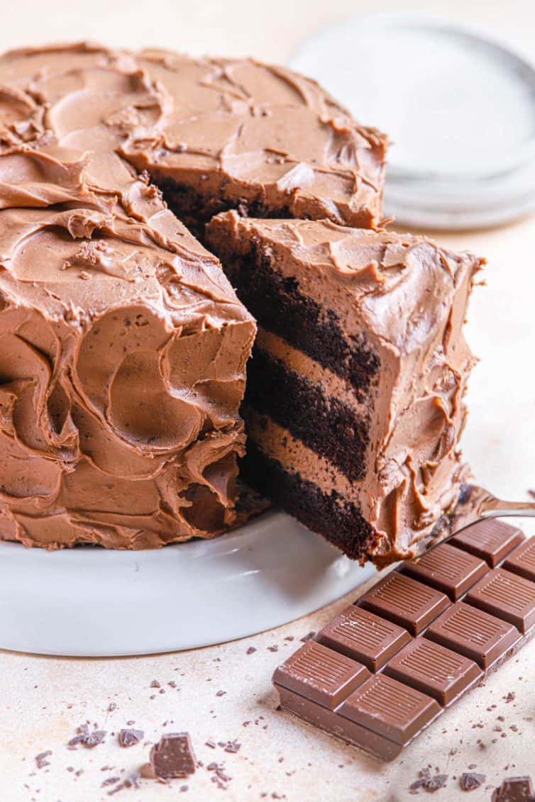 A chocolate cake on a cake stand with a slice of chocolate cake being removed and a chocolate bar in the bottom right.