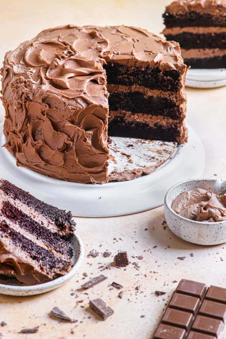 A chocolate cake on a white cake stand with chocolate cake slices in the back and front of the chocolate cake.