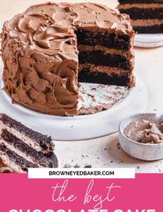 A chocolate cake on a white cake stand with extra frosting in the front right and cake slices in the front left and back right.