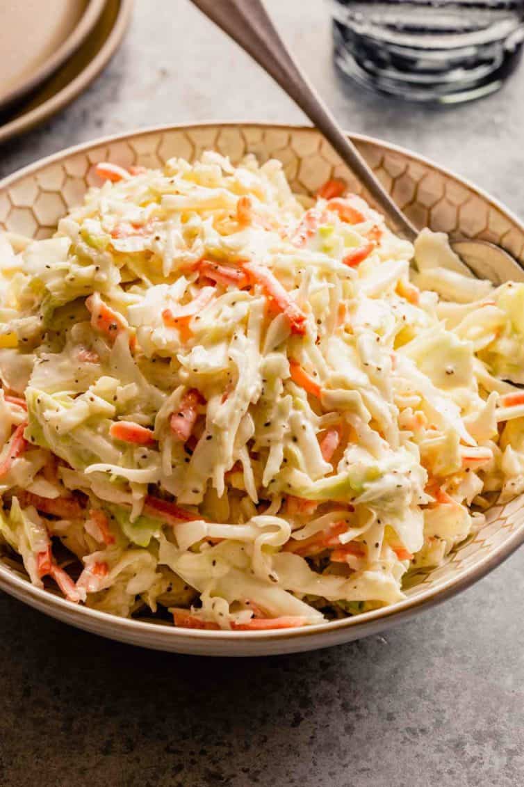 A bowl of creamy coleslaw with a silver spoon on a grey counter.