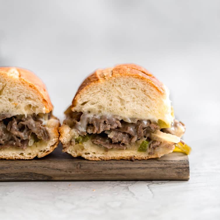 Two philly cheesesteak sandwiches on a wooden cutting board with the insides exposed showing the cheesy meat filling.