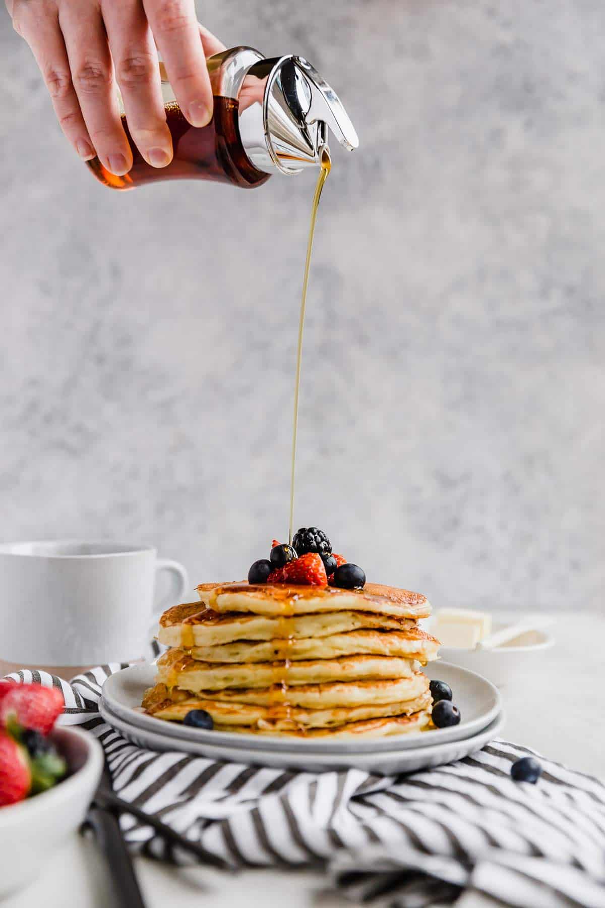A hand pouring maple syrup over the stack of buttermilk pancaked topped with fresh berries on a blue and white striped towel.