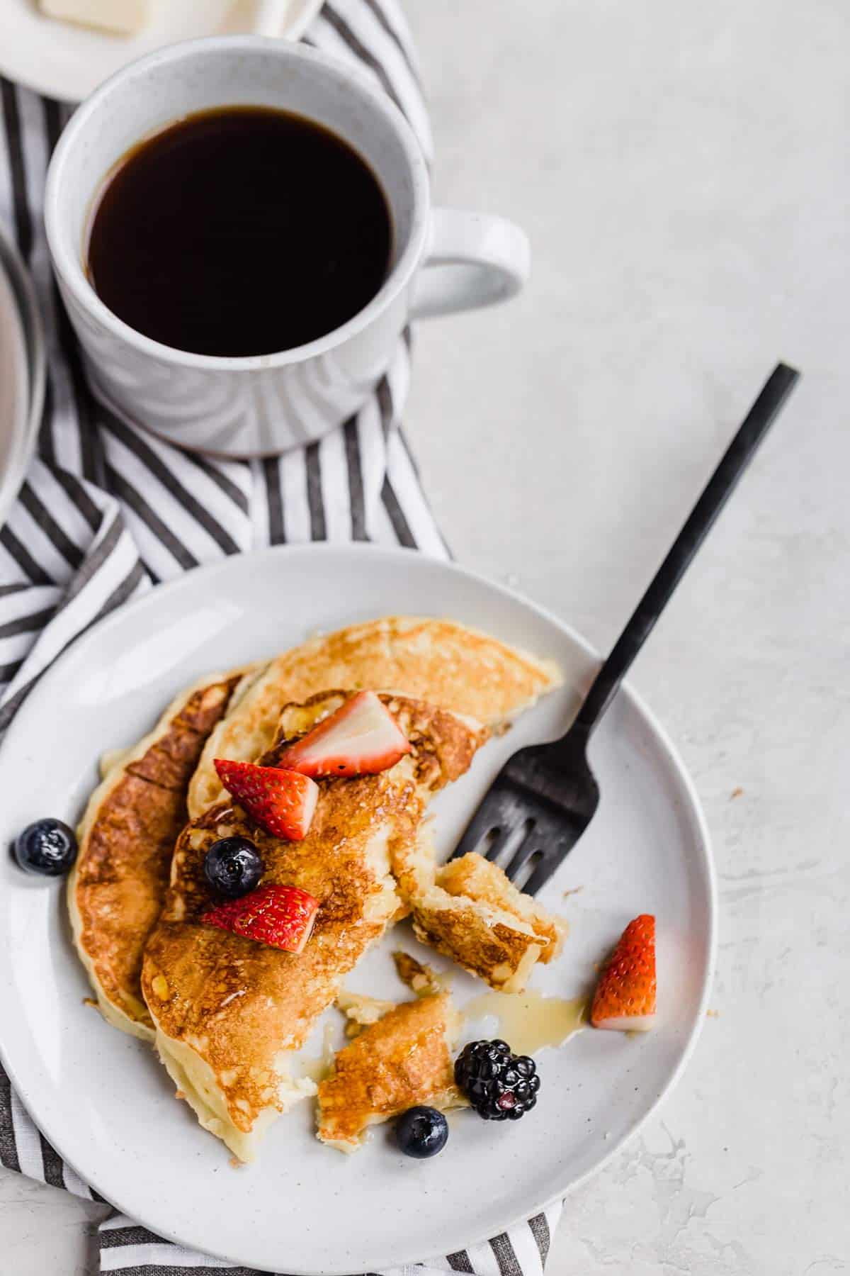 A top down photo of buttermilk pancakes with bites take out and a fork in the top right of the plate with a coffee cup in the top left of the photo with a blue and white striped towel below.