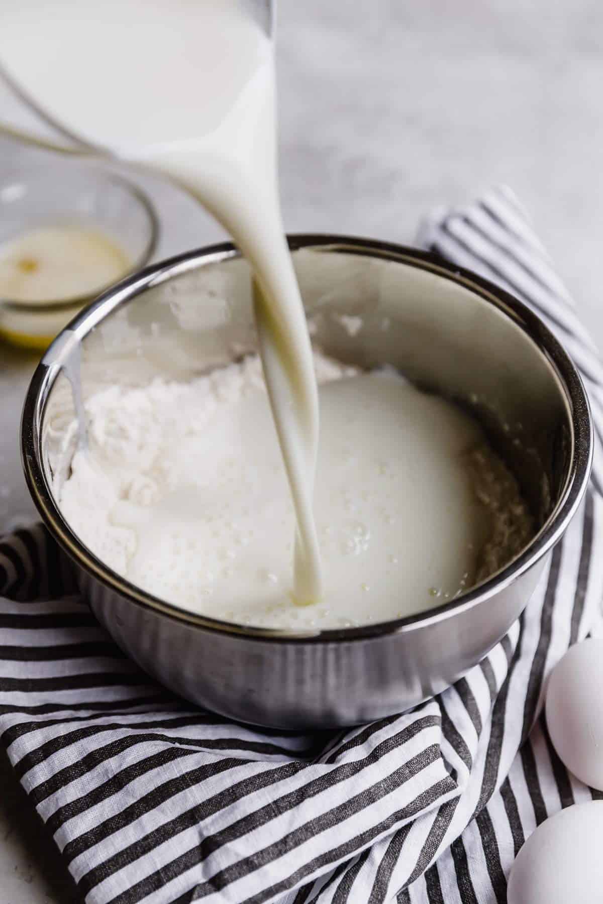 Milk being poured into a bowl of dry ingredients on a counter with a blue and white striped towel.