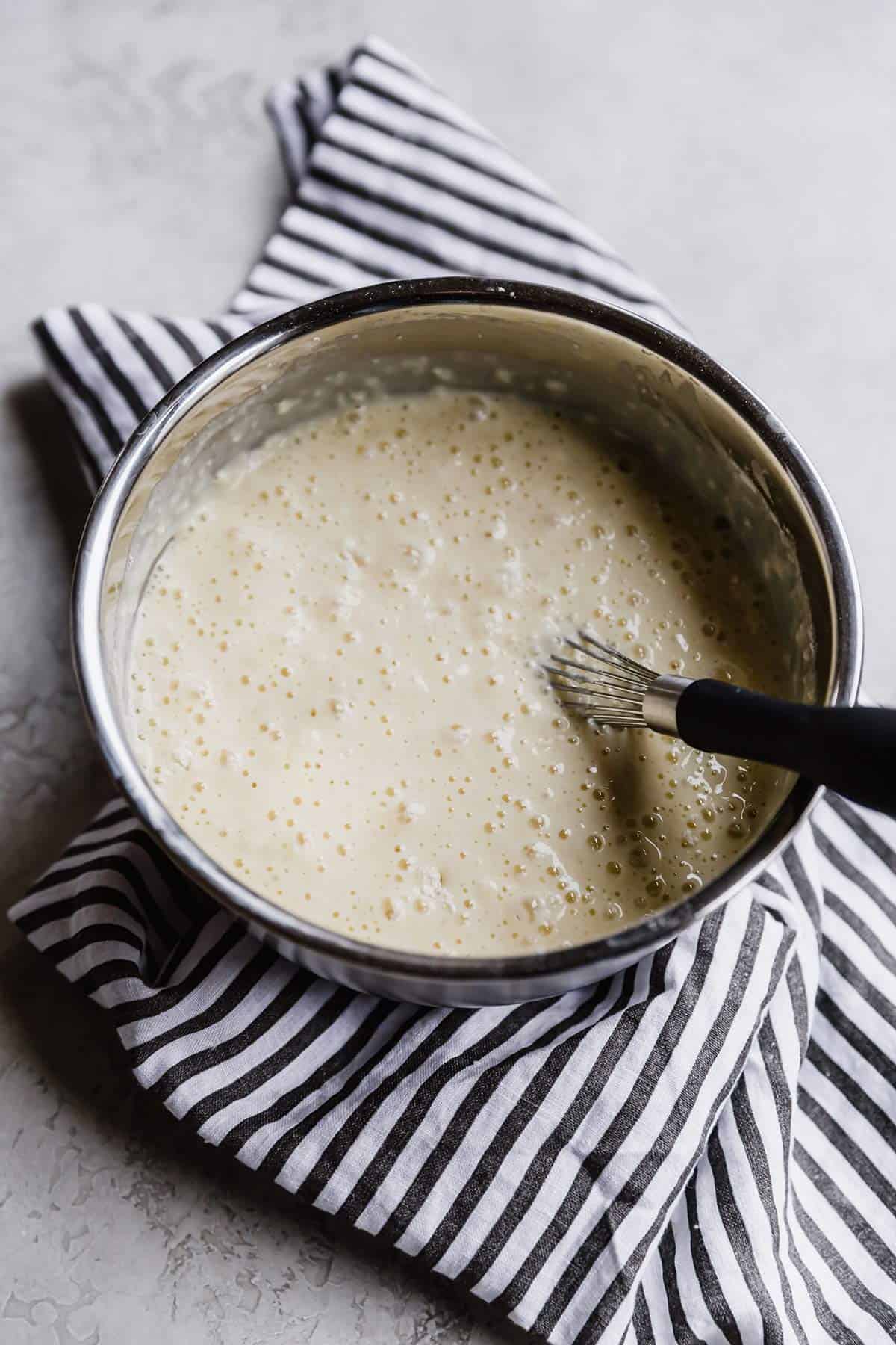 A silver bowl with a wire whisk in the right side of the bowl filled with pancake batter.