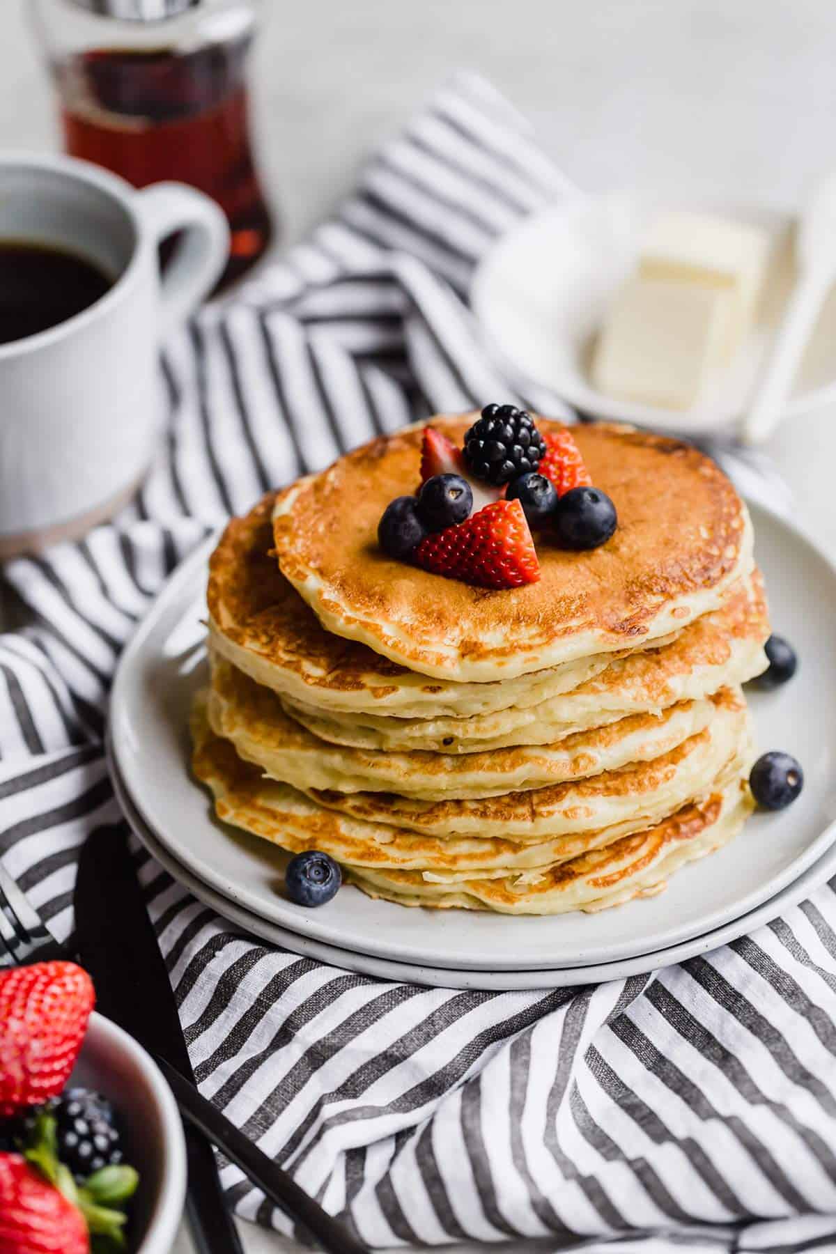 A stack of fluffy buttermilk pancakes topped with fresh berries on a white plate on a white and blue towel with syrup and butter in the back and a small bowl of strawberries in the bottom left corner.