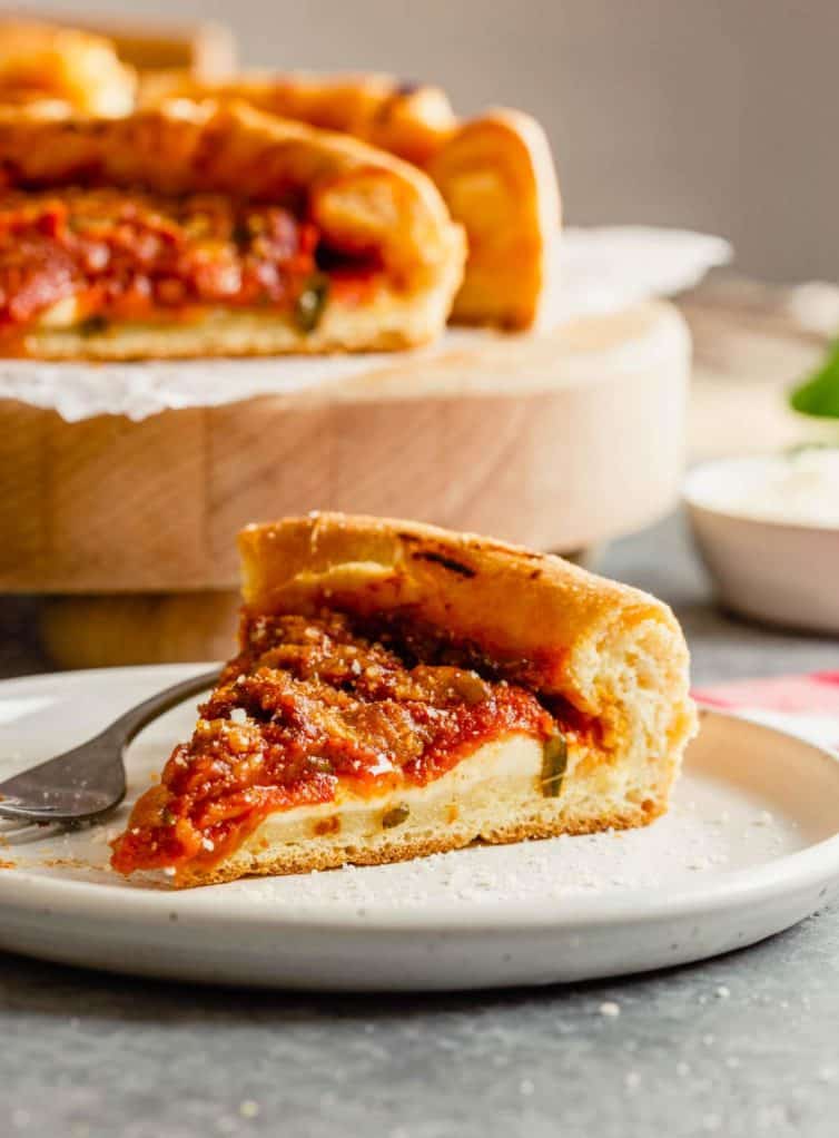 A slice of Chicago-style deep dish pizza on a white plate with a pizza in the back.