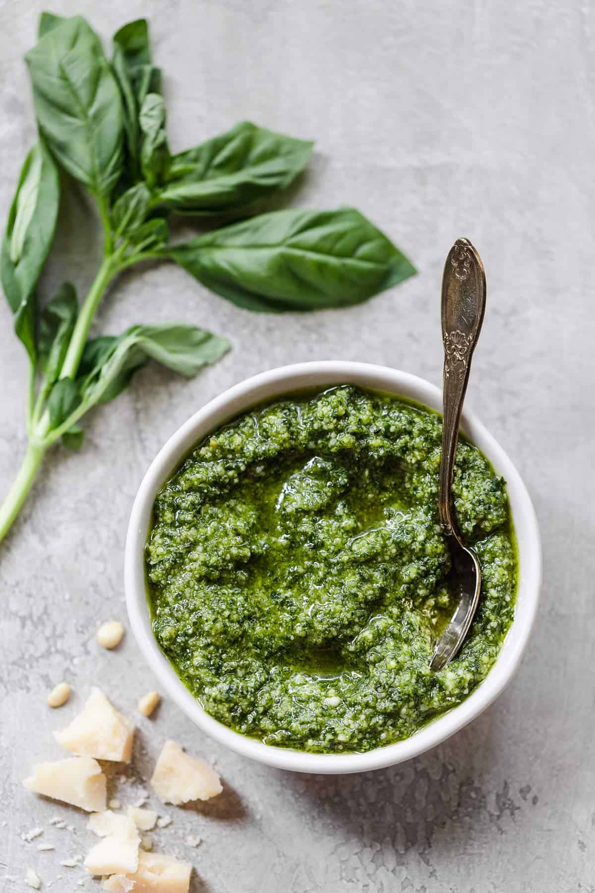 A white bowl with homemade pesto with a spoon in the right side of the bowl and fresh basil on the counter to the top left.