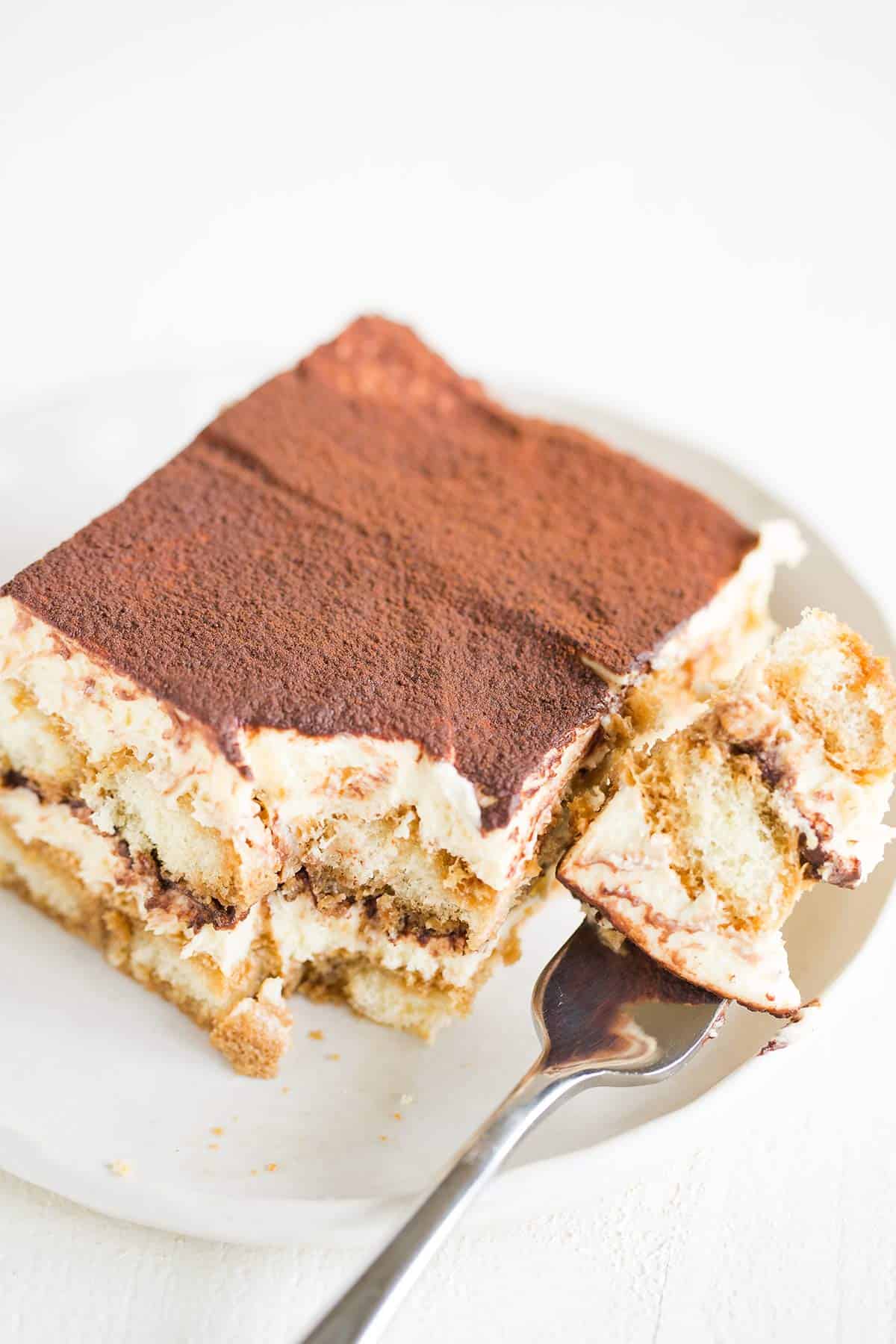 A square slice of tiramisu on a white rimmed plate with a fork full of tiramisu to the right.