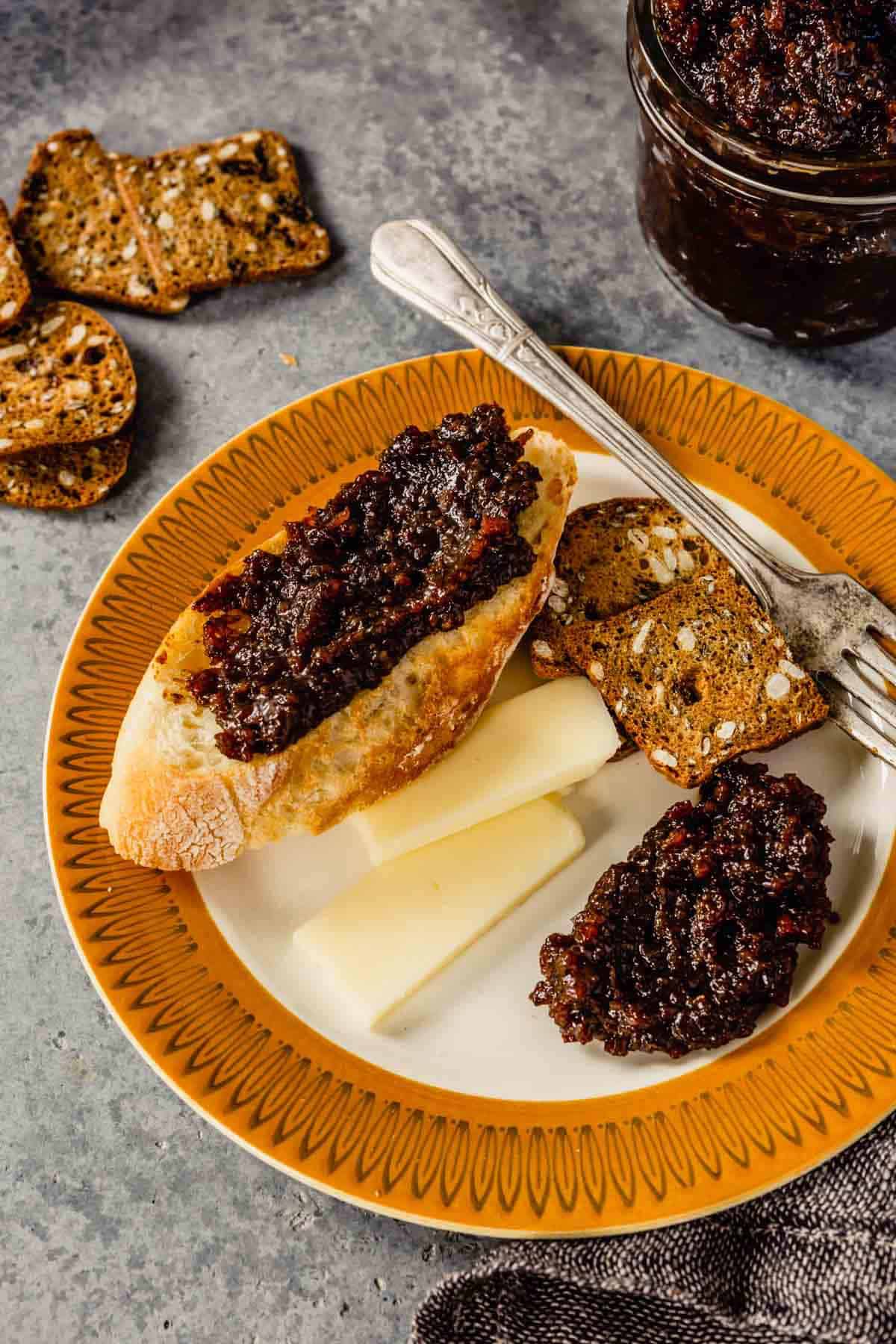An orange rimmed plate with a silver fork and bread with bacon jam, some cheese, and a few crackers on the plate.