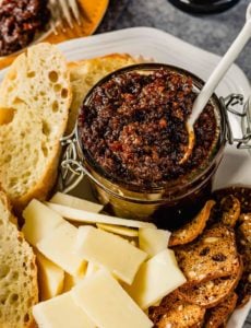 A glass jar of bacon jam with a spoon on a white plate with cheese in front and crackers to the left.