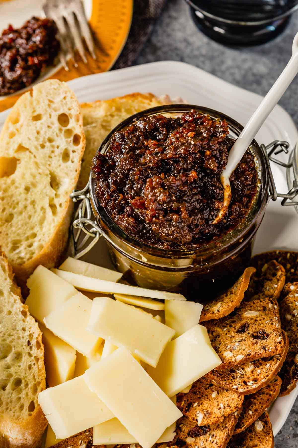 A glass jar of bacon jam with a spoon on a white plate with cheese in front and crackers to the left.