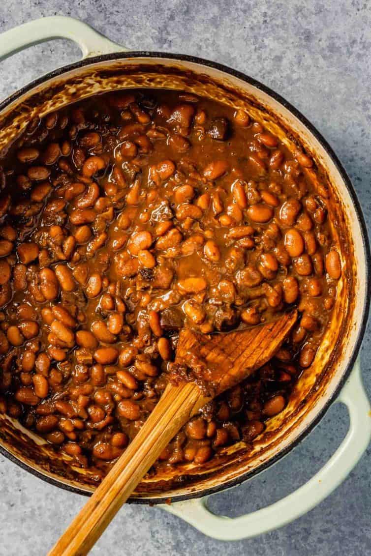 A white dutch oven with Boston baked beans and a wooden spoon to the left.