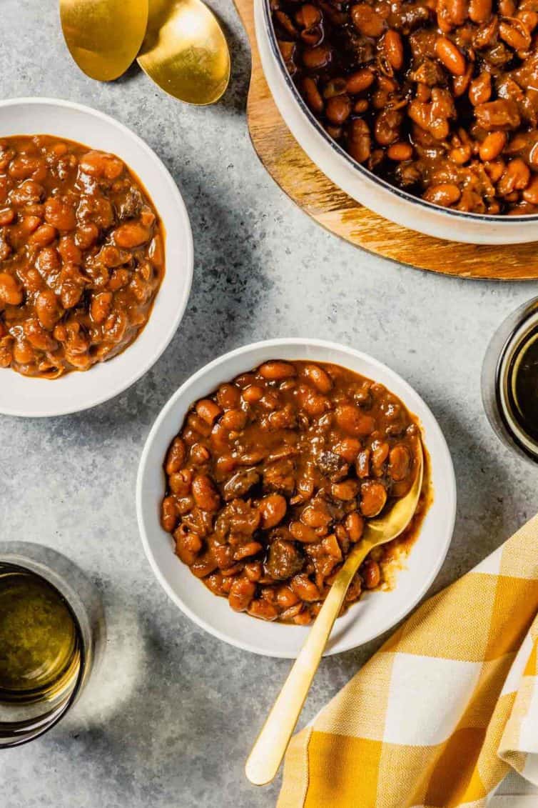 A white bowl with baked beans and a spoon on a grey counter with beans in another white bowl to the top left and top right.