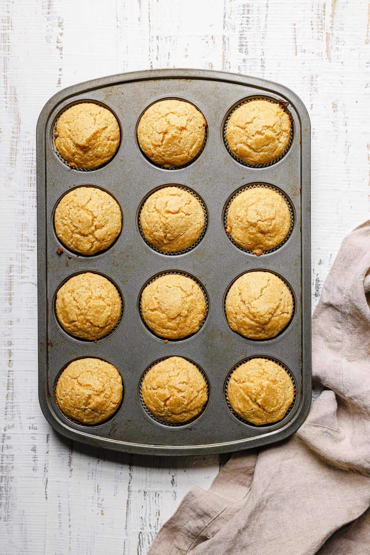 A silver muffin tin with cornbread muffins on a white counter with a light beige dish towel to the left.