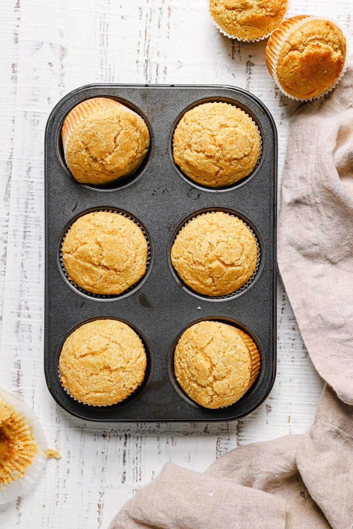 Top down photo of a muffin tin with 6 cornbread muffins on a white counter with two muffins in the top right and a towel in the bottom right.