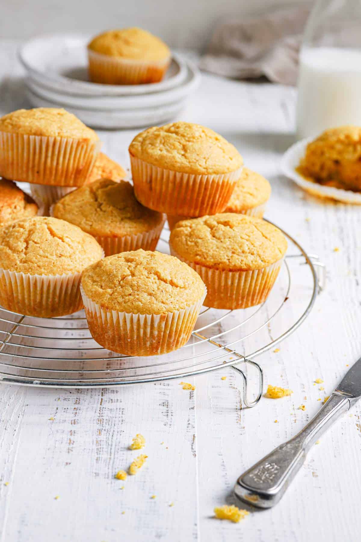 A wire cooling rack with cornbread muffins in a stack with a stack of plates in the back topped with a cornbread muffin and a milk jug to the right.