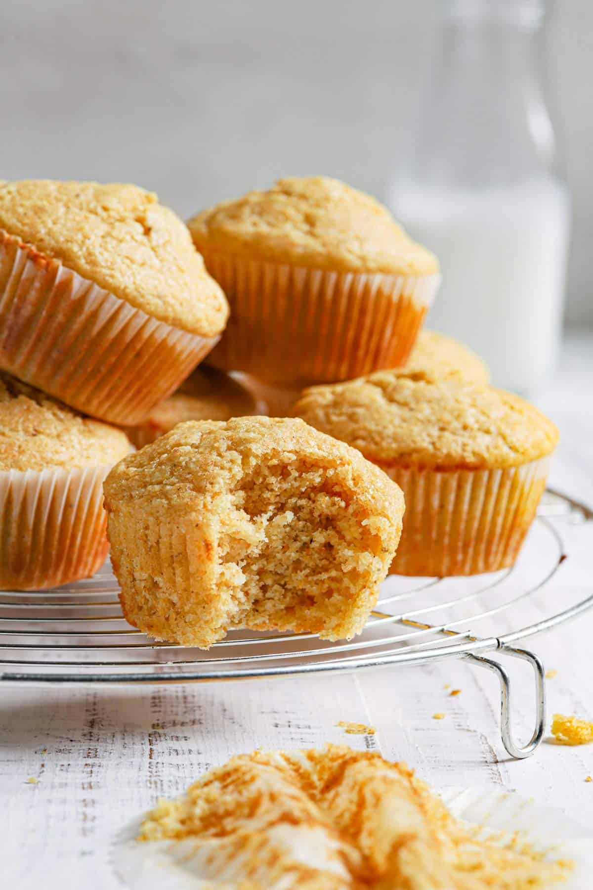 Fresh cornbread muffins on a wire rack with the front muffin missing a bite and a jug of milk in the back.