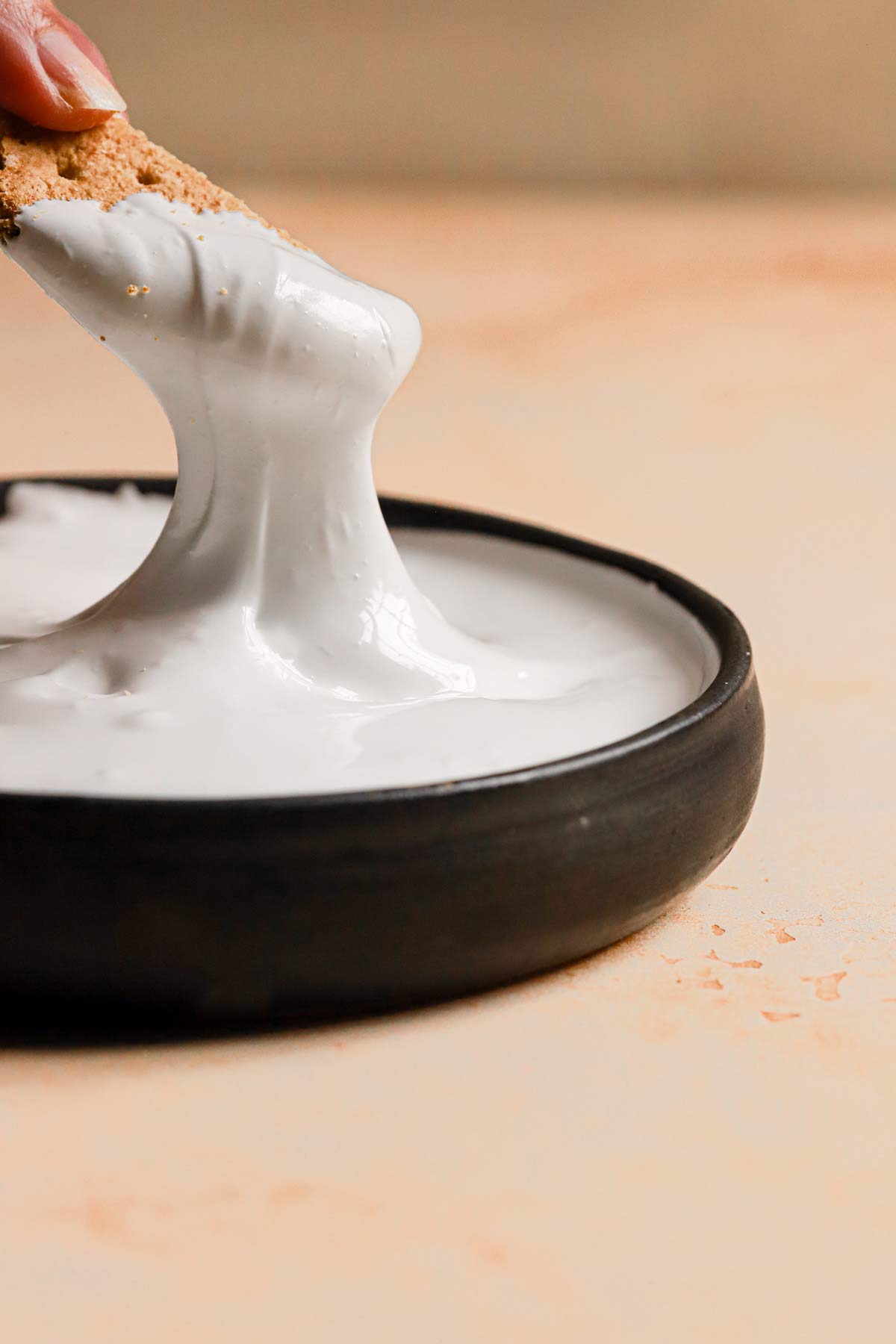 A black bowl on a peach counter with a graham cracker dipping into the marshmallow cream.