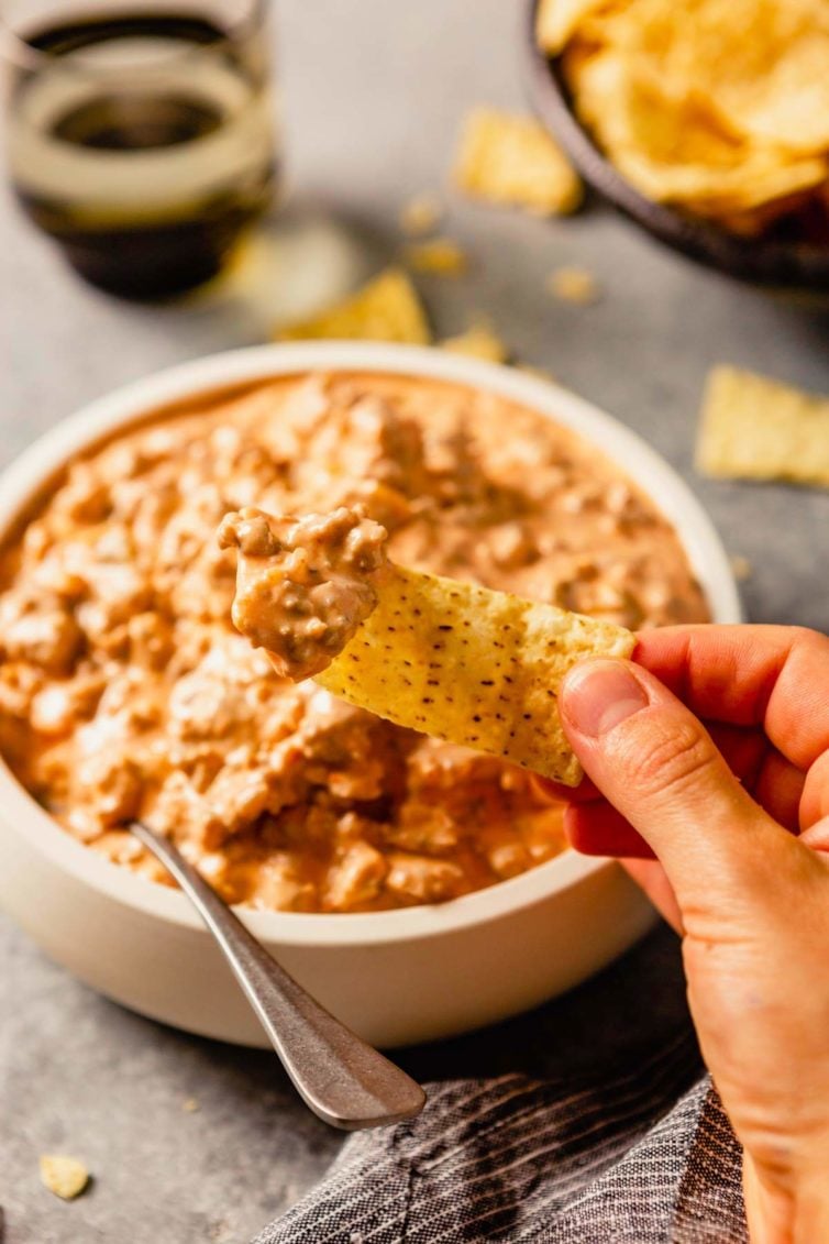 A white bowl with sausage dip and a hand holding a chip in front after dipping in the sausage dip.